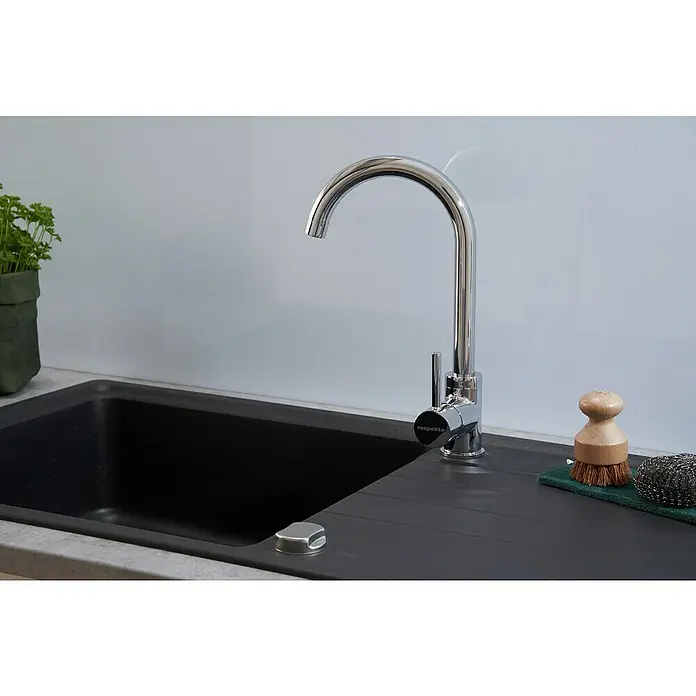 Respekta Einbauspüle Columbia - 100 x 50 cm, Schwarz - Ein hochglanzpolierter, verchromter Wasserhahn der Marke Respekta steht neben einer Spüle. Auf der Arbeitsplatte befindet sich eine Bürste mit Spülschwamm.