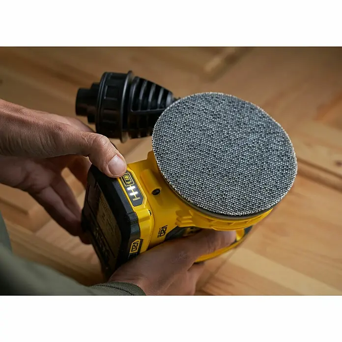 Stanley FatMax Akku-Exzenterschleifer SFMCW220D1S-QW - 18 V, 1 Akku, Durchmesser Schleifteller: 125 mm - Nahaufnahme eines gelben DeWalt-Schleifers mit einem Akku, der von zwei Händen gehalten wird und auf einem Holzboden steht.