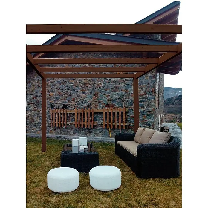 Pérgola estándar Green - L x An x Al: 400 x 300 x 250 cm, Marrón - Zona de relax al aire libre con sofá de ratán negro, mesa auxiliar y dos pufs blancos bajo una pérgola de madera frente a un muro de piedra.