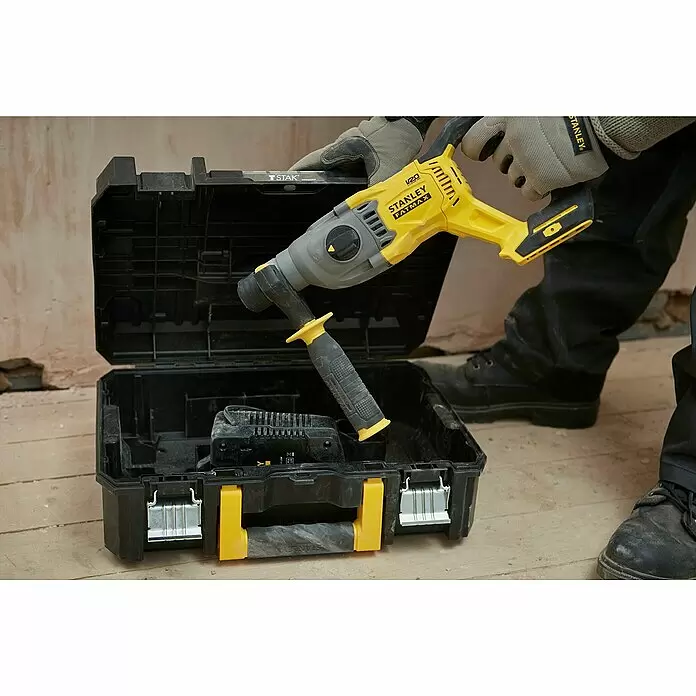 Stanley FatMax Akku-Kombihammer SFMCH900B-XJ - 18 V, Ohne Akku, 2 J - Eine Person in Arbeitshandschuhen legt einen gelb-grauen Stanley Fatmax V20 Bohrhammer in eine schwarze Werkzeugbox, die ein Akku-Ladegerät enthält.