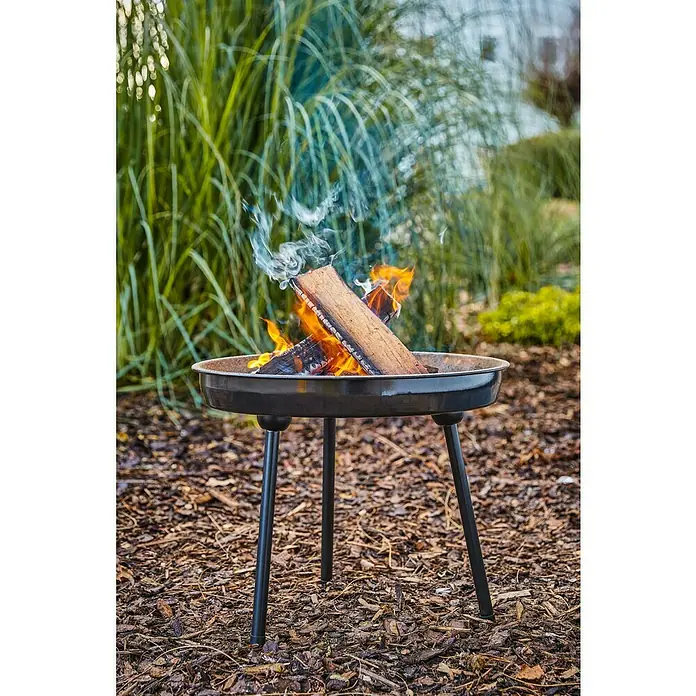 Grillstar Schwenkgrill Louisiana - Durchmesser Grillfläche: 47 cm, Höhe: 160 cm, Schwarz - Das Bild zeigt eine Feuerschale mit brennendem Holz, die auf einem Beet mit Rindenmulch steht. Im Hintergrund sind Gräser und Büsche zu sehen.