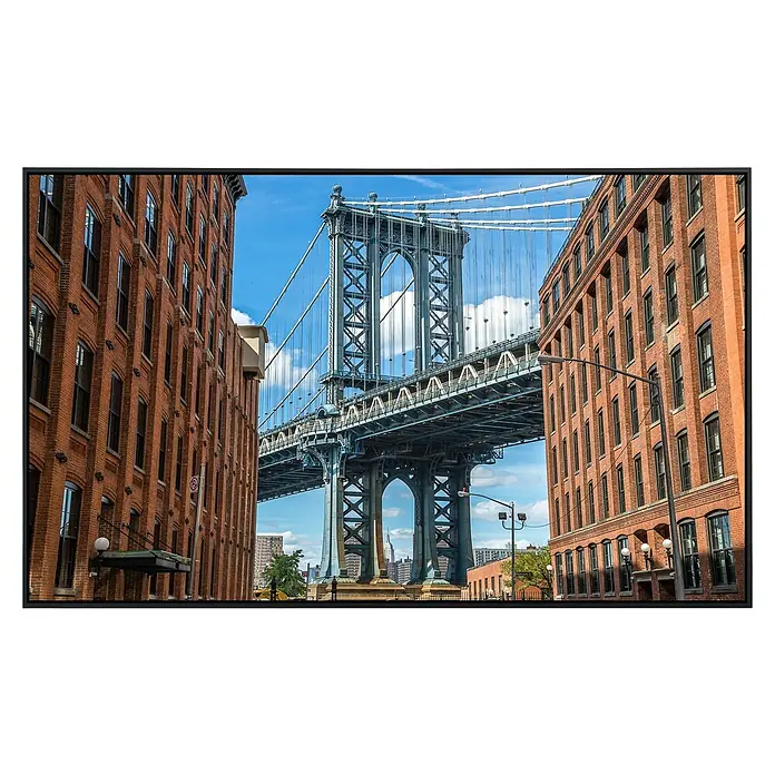 Papermoon Infrarot-Bildheizkörper Brooklyn Dumbo Bereich - 60 cm x 500 mm, 350 W - Ein TV-Gerät zeigt ein Foto der Manhattan Bridge, die zwischen zwei Backsteingebäuden hindurchführt, unter blauem Himmel.