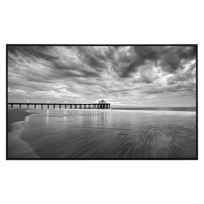 Papermoon Infrarot-Bildheizkörper Manhattan Beach Pier - 90 x 50 cm, 450 W - Schwarz-Weiß-Fotografie einer Seebrücke unter einem bewölkten Himmel, eingefasst in einem eleganten schwarzen Rahmen. Die glatte Wasseroberfläche reflektiert das Licht.