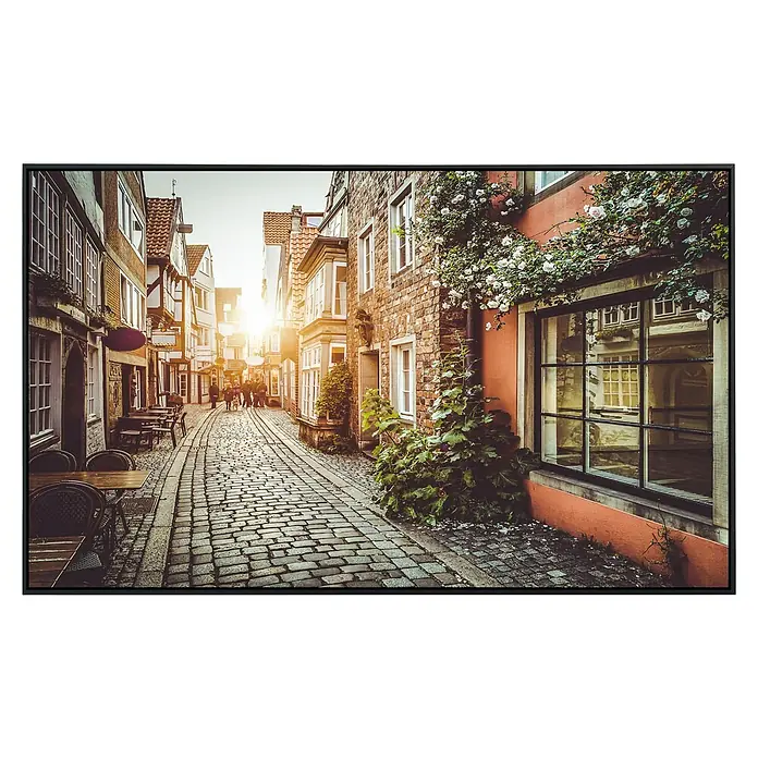 Papermoon Infrarot-Bildheizkörper Altstadt in Belgien - 90 x 50 cm, 450 W - Gemälde mit Ansicht einer malerischen Gasse mit Kopfsteinpflaster, gesäumt von historischen Gebäuden und üppigem Grün in warmen Farben.