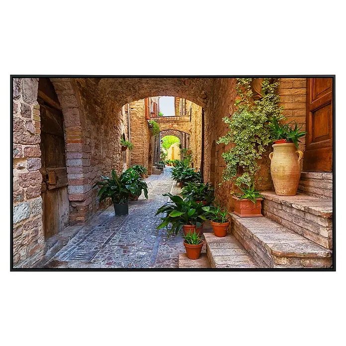 Papermoon Infrarot-Bildheizkörper Antike Stadt Spello Umbrien - 120 x 60 cm, 1.000 W - Ein schmaler, gepflasterter Weg mit blühenden Pflanzen und antiken architektonischen Details, gerahmt von einem schwarzen Rahmen.