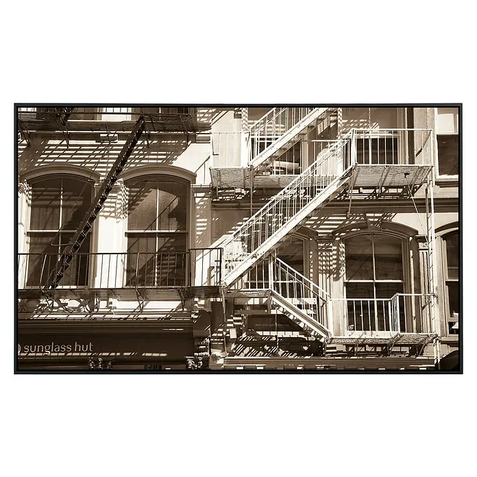 Papermoon Infrarot-Bildheizkörper Soho New York - 100 x 60 cm, 600 W - Fassade eines Gebäudes mit einer markanten Außentreppe, die sich vor den Fenstern schlängelt. Im Erdgeschoss befindet sich ein "Sunglass Hut" Geschäft.