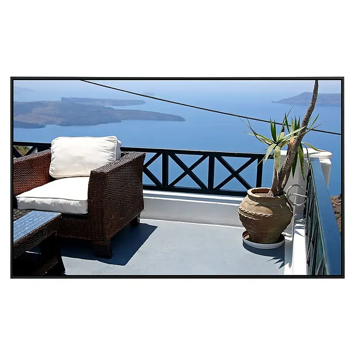 Papermoon Infrarot-Bildheizkörper Santorini Inselblick - 90 x 50 cm, 450 W - Einladender Außenbereich mit Korbsessel und Meerblick. Der Sessel mit weißen Kissen lädt zum Verweilen ein, während die Aussicht begeistert.