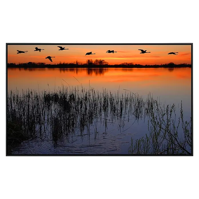 Papermoon Infrarot-Bildheizkörper Abenddämmerung am See - 100 x 60 cm, 800 W - Malerisches Landschaftsbild eines Sees mit Schilf und fliegenden Schwänen bei Sonnenuntergang, gerahmt in einem eleganten schwarzen Rahmen.