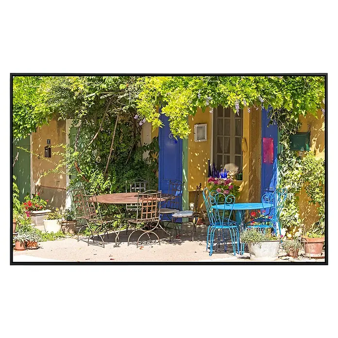 Papermoon Infrarot-Bildheizkörper Provence Cafe Shop - 100 x 60 cm, 800 W - Ein sonniger Innenhof mit Tischen und Stühlen in verschiedenen Farben vor einem charmanten Haus mit bunten Details.