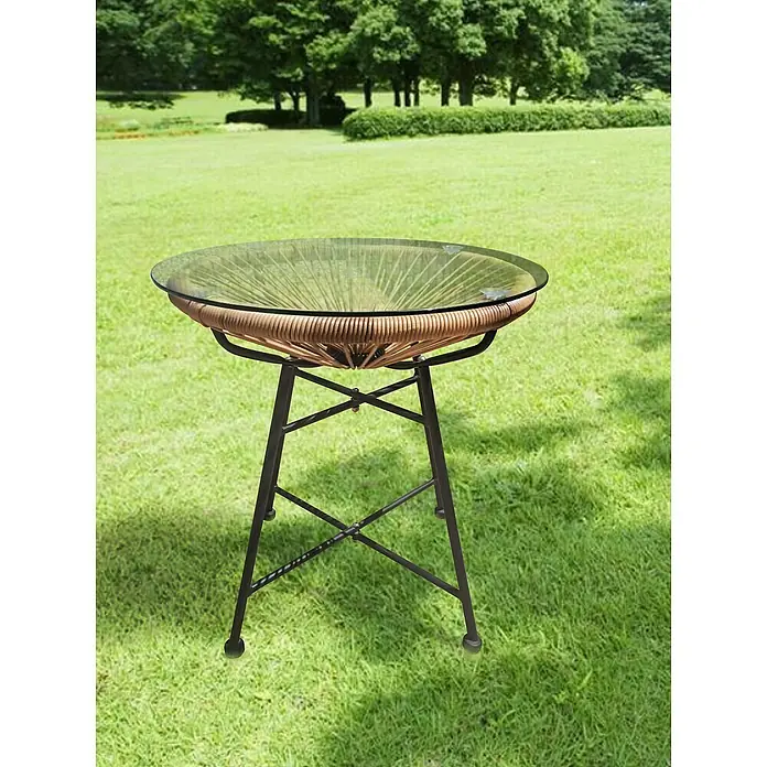 Mesa auxiliar de jardín redonda Fidji - Ø x Al: 45 x 45 cm, Marrón/Negro - Una mesa redonda de jardín con tapa de vidrio y tejido de ratán sobre un césped verde. La estructura de la mesa es de metal oscuro.