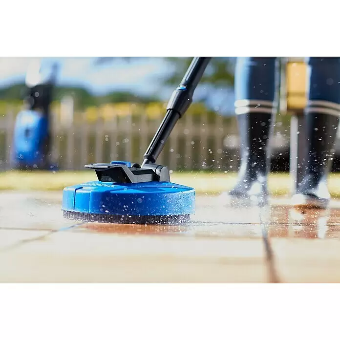 Nilfisk Hochdruckreiniger C 135.7-6 Garden X-tra - 1.500 W, Max. Druck: 135 bar, Fördermenge: 460 l/h - Blau-schwarzer Hochdruckreiniger-Flächenreiniger reinigt aktiv eine Terrasse, Wasser spritzt. Person in Stiefeln verschwommen im Hintergrund.