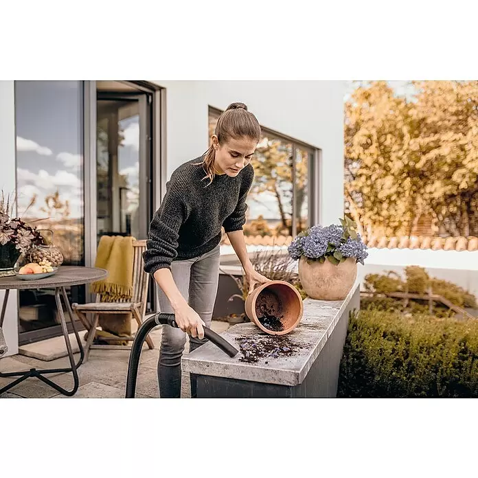 Kärcher Elektro-Nass- & Trockensauger WD 3 S V-17/4/20 - 1.000 W, Behältervolumen: 17 l - Frau saugt verschüttete Erde und Blüten von einem Topf auf einer Terrassenkante mit einem schwarzen Staubsaugerschlauch auf.
