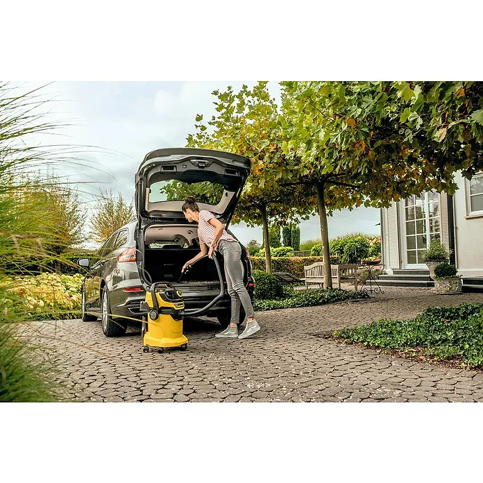 Kärcher Elektro-Nass- & Trockensauger WD 5 V-25/5/22 - 1.100 W, Behältervolumen: 25 l - Eine Frau benutzt einen Kärcher Mehrzwecksauger zum Reinigen des Kofferraums ihres Autos. Der gelbe Sauger steht auf dem gepflasterten Untergrund.
