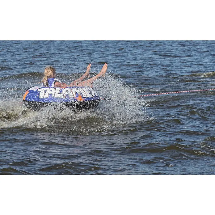 Talamex Funtube Fire - Durchmesser: 137 cm, Geeignet für: 1 Person, Schnellbefestigung - Ein Mädchen saust auf einem blauen TALAME-Reifen hinter einem Boot her, umgeben von spritzendem Wasser.