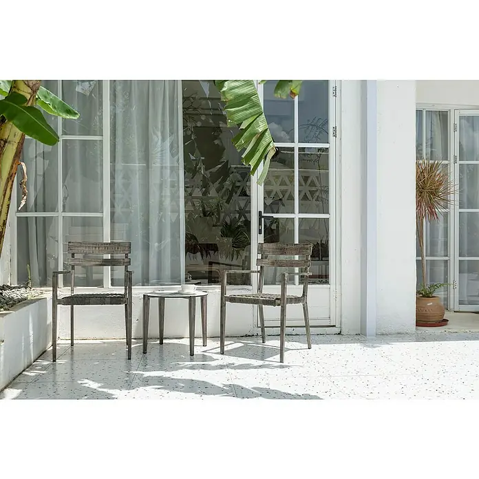 Silla de jardín Josefina - Bicolor gris, Apilable - Dos sillas de madera grises y una mesa se encuentran en un patio frente a una puerta blanca, rodeadas de exuberante vegetación.