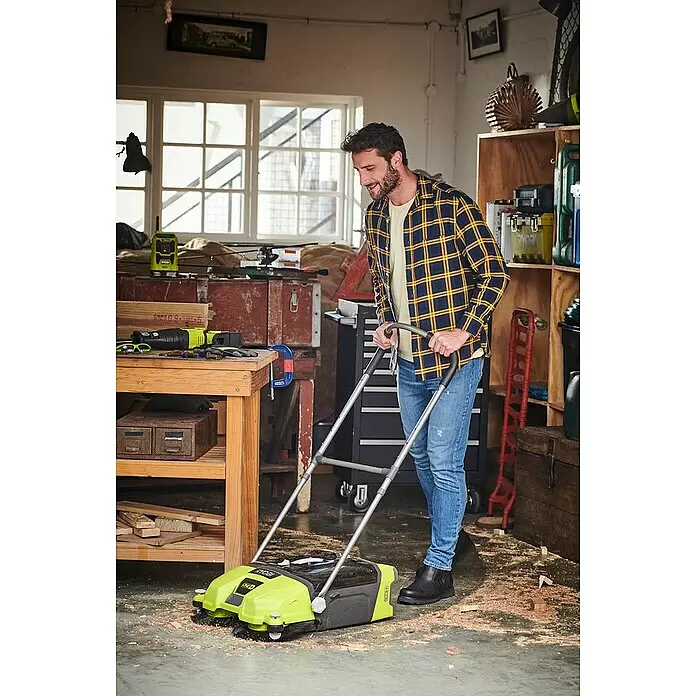 Ryobi ONE+ Akku-Handkehrmaschine R18SW3-120S - 18 V, 17 l, 1 Akku - Ein Mann lächelt, während er eine grün-schwarze Ryobi Kehrmaschine benutzt, um Sägemehl und Schmutz vom Werkstattboden zu entfernen.