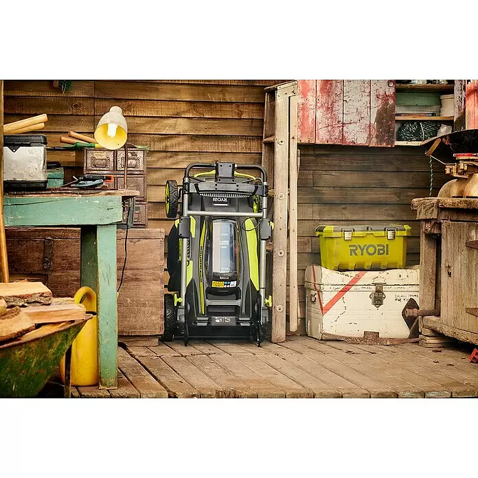Ryobi Akku-Rasenmäher RY36LMX51A-160 - 36 V, 1 Akku, 6 Ah, Schnittbreite: 51 cm - Ryobi Rasenmäher, schwarz und lindgrün, hochgeklappt zur kompakten Lagerung in einem rustikalen Holzschuppen.