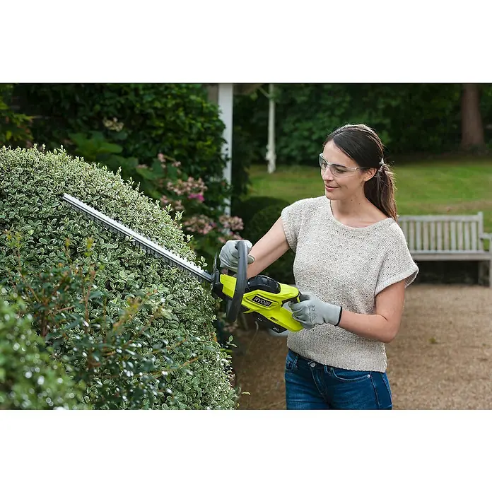 Ryobi ONE+ Akku-Heckenschere RHT184520 - 18 V, Li-Ionen, 1 Akku, Schnittlänge: 45 cm - Frau, die eine Ryobi-Heckenschere benutzt, um im Garten einen Busch zu schneiden. Sie trägt Schutzkleidung und arbeitet sorgfältig.
