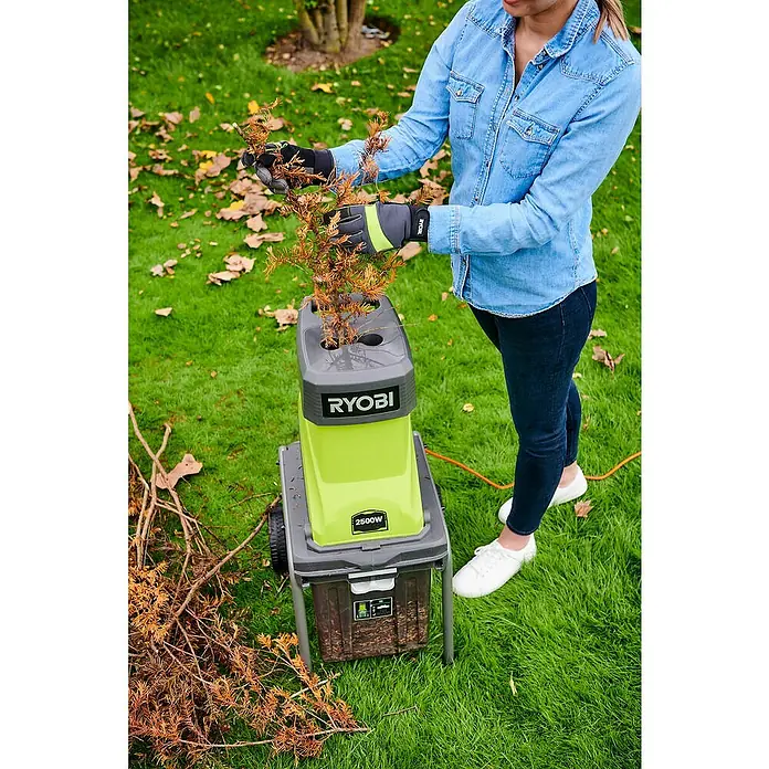 Ryobi Elektro-Häcksler RSH2545B - Max. Aststärke: 4,5 cm, 2.500 W - Eine Frau benutzt einen Ryobi-Häcksler im Garten, um Äste zu zerkleinern. Der Häcksler ist grün und grau und steht auf dem Rasen.