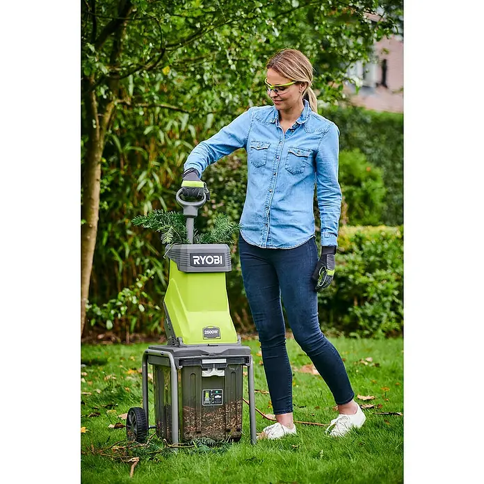 Ryobi Elektro-Häcksler RSH2545B - Max. Aststärke: 4,5 cm, 2.500 W - Eine Frau bedient einen Ryobi 2500W Häcksler im Garten, umgeben von Grün. Der Häcksler steht auf einem Rahmen mit Rädern für Mobilität.