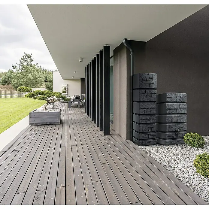 Regenspeicher Tower Stone - 500 l, Anthrazit - Aufnahme von zwei dunklen, steinartigen Regenwassertanks auf einer Holzterrasse neben einem modernen Haus.