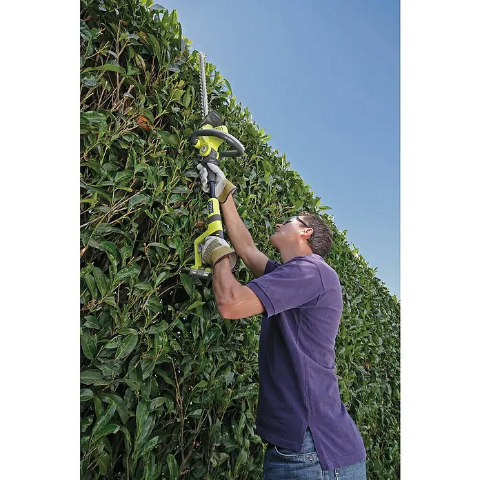 Ryobi ONE+ Maschinenpaket - Akku-Heckenschere OHT1850X,18V, 4 Ah Akku & 2A Ladegerät - Ein Mann, der mit einer Ryobi-Heckenschere eine hohe grüne Hecke schneidet, wobei der Fokus auf dem Gartengerät liegt.
