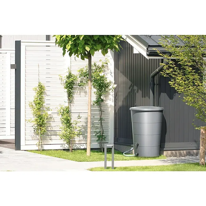 Regentonne Smooth - 160 l, Hellgrau - Eine graue Regentonne steht neben einem Haus mit einem Fallrohr. Die Tonne sammelt Regenwasser für die Gartenarbeit und ist umweltfreundlich.
