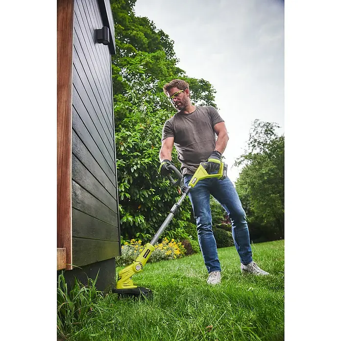 Ryobi ONE+ Akku-Rasentrimmer OLT1832 - 18 V, Li-Ionen, Ohne Akku - Ein Mann benutzt einen Ryobi Rasentrimmer, um Gras an einer dunklen Gebäude-Fassade zu trimmen. Er trägt Schutzkleidung und arbeitet im Garten.