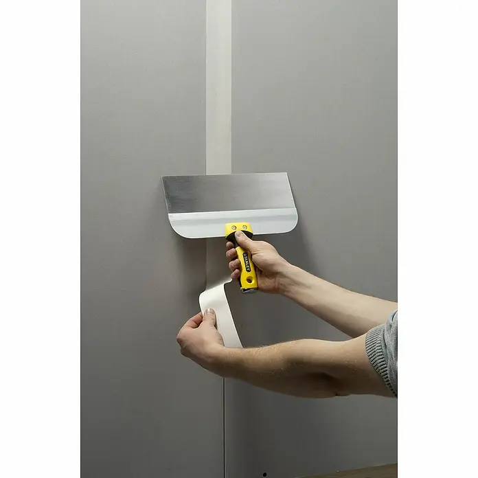 Stanley Breitspachtel STHT0-05771 - Stahl rostfrei, 250 mm - Die Hand eines Mannes verwendet eine Spachtel zum Anbringen von Malerkrepp an einer Wand.