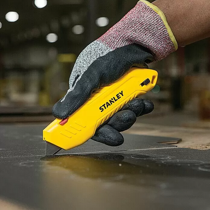 Stanley Sicherheitsmesser STHT10368-0 - Trapezklinge - Eine behandschuhte Hand hält ein gelbes STANLEY Cuttermesser und schneidet in eine dunkle Oberfläche.