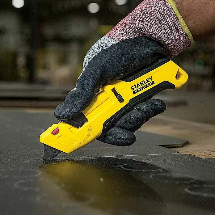 Stanley FatMax Sicherheitsmesser FMHT10365-0 - Trapezklinge - Eine behandschuhte Hand hält ein gelbes Stanley FatMax Cuttermesser und schneidet ein dunkles Material.