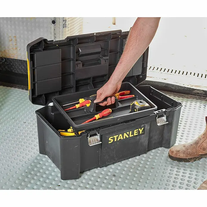 Stanley Werkzeugkasten ESSENTIAL™ STST82976-1B x L: 67 x 28 cm, Kunststoff - Ein schwarzer Werkzeugkasten von Stanley wird von einer Hand geöffnet, die eine Werkzeugablage mit Schraubenziehern und einem Maßband hält.