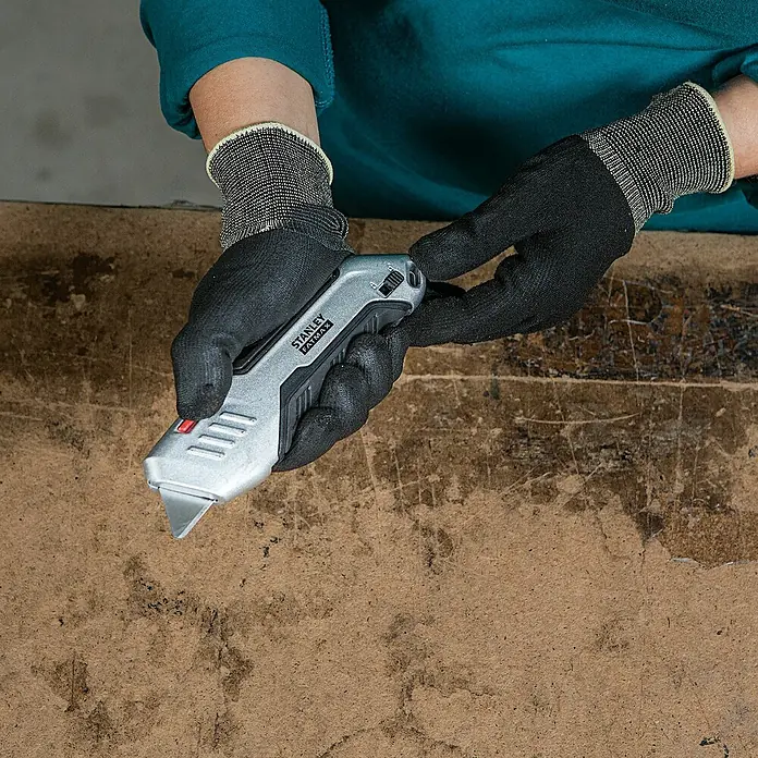 Stanley FatMax Sicherheitsmesser FMHT10370-0 - Trapezklinge - Hände mit Schutzhandschuhen halten ein Stanley Fatmax Cuttermesser über einer Holzoberfläche.