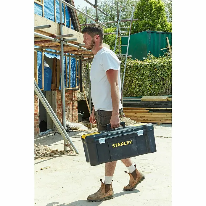 Stanley Werkzeugkasten ESSENTIAL™ STST82976-1 - B x L: 67 x 28 cm, Kunststoff - Ein Bauarbeiter trägt einen schwarzen Stanley Werkzeugkasten. Der Kasten ist robust und ideal für den Transport von Werkzeugen.