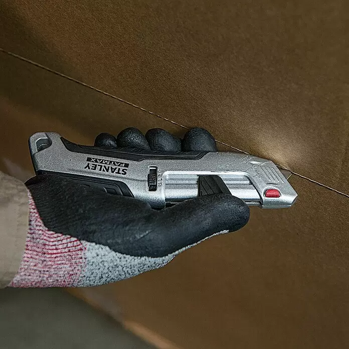 Stanley FatMax Sicherheitsmesser FMHT10367-0 - Trapezklinge - Eine behandschuhte Hand hält ein silbernes Stanley FatMax Cuttermesser, das in einen braunen Karton schneidet.