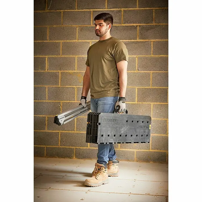 Stanley Werkbank ESSENTAIL™ STST83492-1 - L x B: 85 x 61 cm, Arbeitshöhe: 80 cm, Belastbarkeit: 320 kg, Schwarz/Gelb - Ein Mann trägt eine Stanley Werkbank und eine Stativleiter vor einer Ziegelwand. Ideal für Heimwerker und Profis.