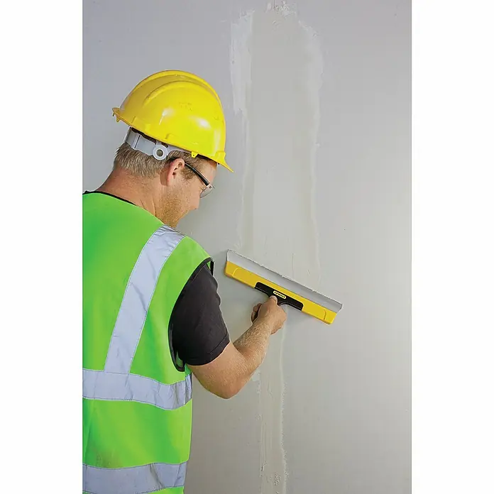 Stanley Flächenspachtel STHT0-05934 - Klingenbreite: 300 mm - Ein Bauarbeiter mit Schutzhelm und Sicherheitsweste glättet eine Wand mit einem gelben Stanley Glättspachtel.