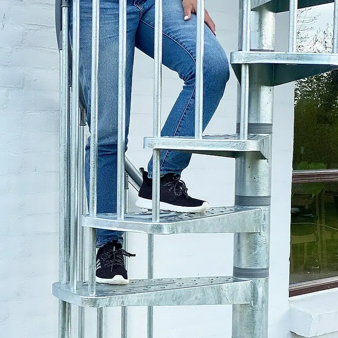 Dolle Außentreppe Gardenspin - Durchmesser: 185 cm, Geschosshöhe: 307,5 cm - 352,5 cm, Lochblechstufen, Silber - Eine Person steigt eine moderne, feuerverzinkte Wendeltreppe im Freien hinauf. Ideal für Garten und Terrasse.