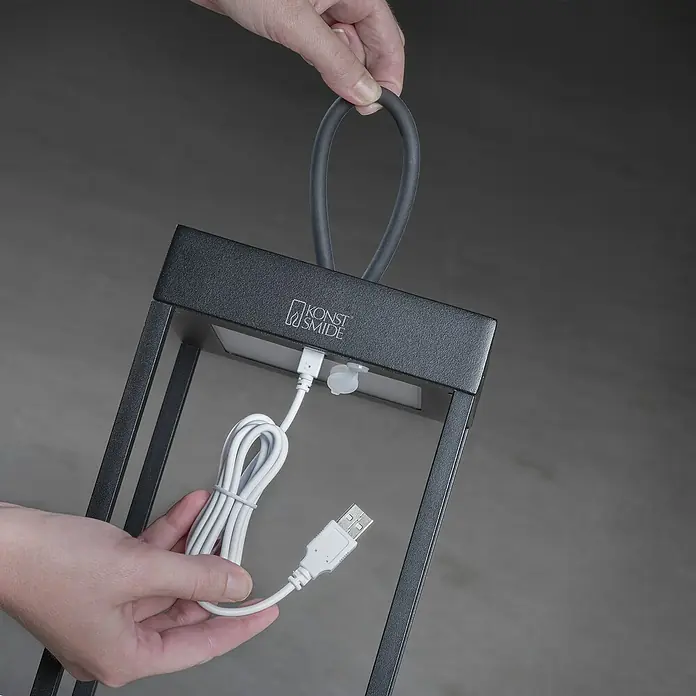 Konstsmide Außenlaterne Ravello -  - Nahaufnahme einer Konst Smide Laterne mit USB-Kabel, die von einer Person gehalten wird, was die einfache Aufladung und Tragbarkeit des Produkts hervorhebt.