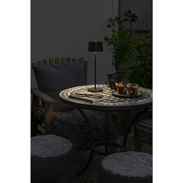 Konstsmide Akku-Tischleuchte Positano - 2,2 W, Schwarz, Warmweiß - Tischszene mit Lampe, Tassen, Muffins und Notizbuch. Die Lampe beleuchtet die Szene und schafft eine gemütliche Atmosphäre.