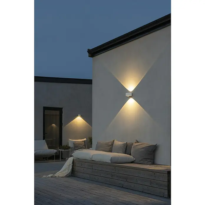 Konstsmide LED-Außenwandleuchte Gala - 6 W, Weiß, IP54 - Eine stilvolle Außenbereichsszene mit einer modernen Wandleuchte, die ein warmes Licht auf eine gemütliche Sitzecke wirft.