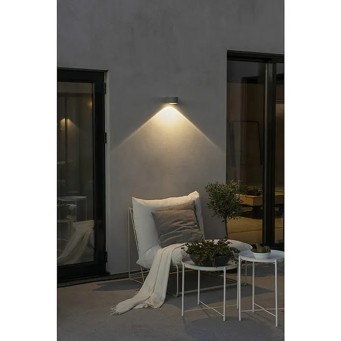 Konstsmide LED-Außenwandleuchte Gala - 6 W, 15,5 x 11 x 6,5 cm, Anthrazit, IP54 - Stilvolle Außenwandleuchte illuminiert eine gemütliche Terrasse mit Sessel, Beistelltischen und dekorativen Pflanzen für eine entspannende Atmosphäre.
