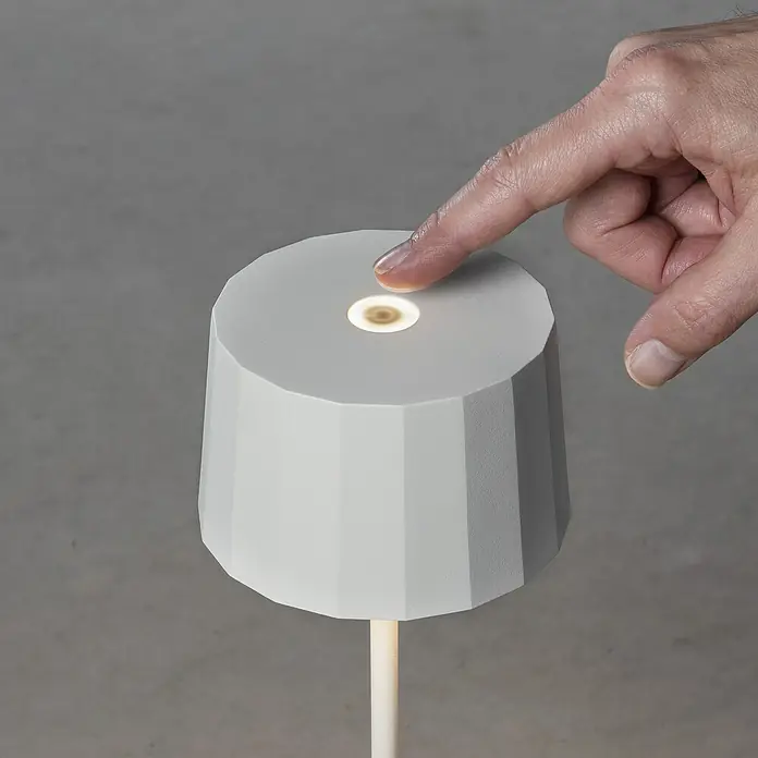 Konstsmide Akku-Tischleuchte Positano - 2,2 W, Weiß, Warmweiß - Ein minimalistisches, modernes Lichtobjekt: Eine Hand tippt auf die Oberseite einer grauen, facettierten Lampe, um sie zu aktivieren.