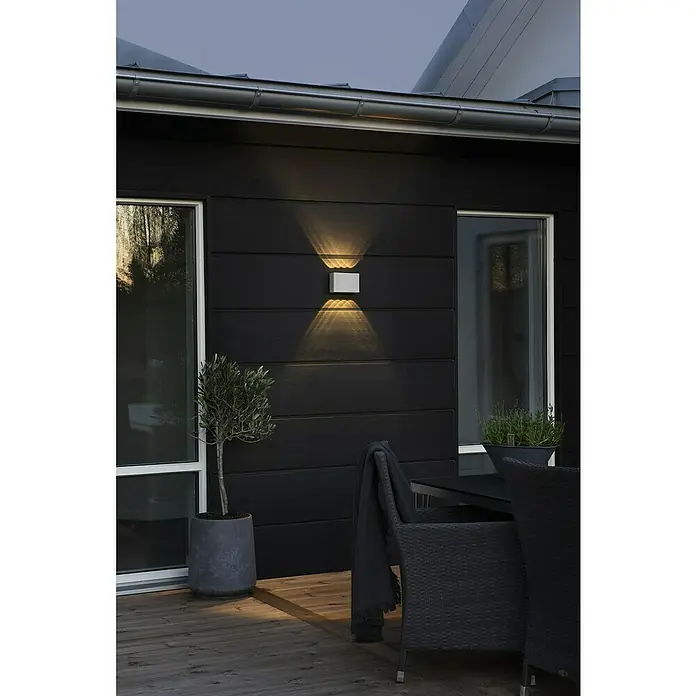 Konstsmide LED-Außenwandleuchte Chieri - 8 W, 6 x 17 x 11 mm, Weiß, IP54 - Eine moderne, silberne Außenleuchte strahlt warmes Licht an einer dunklen Hauswand ab und erhellt stilvoll den Außenbereich.