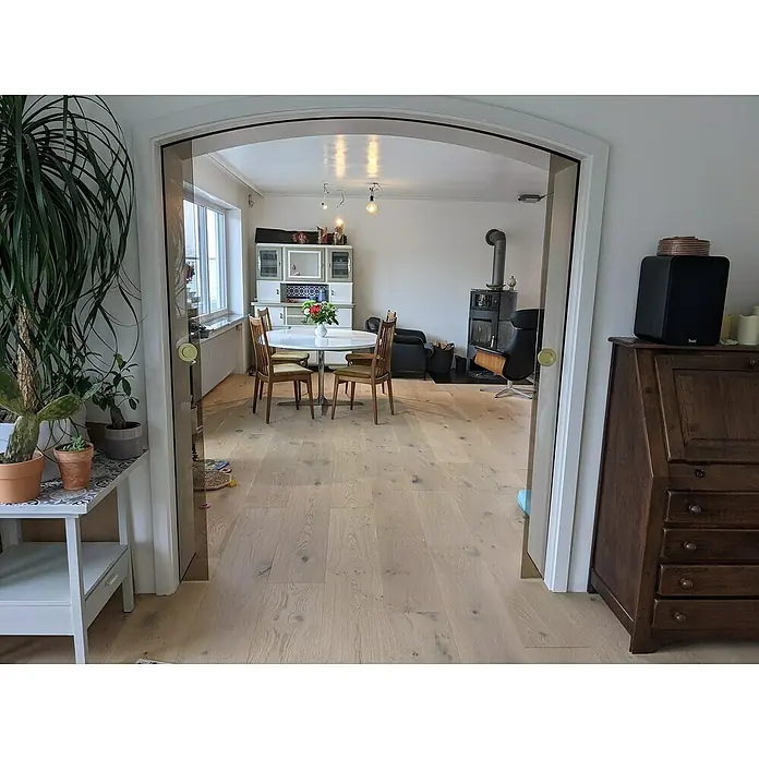 Parkettmuster Eiche Inntal rustikal - 300 x 190 x 14 mm, Landhausdiele - Blick durch einen eleganten Türrahmen auf einen hellen Essbereich mit Holzboden, umgeben von stilvollen Möbeln und dekorativen Pflanzen.