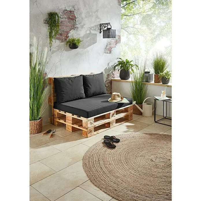 Rückenkissen Virginia - 45 x 60 x 12 cm, Anthrazit - Palettensofa mit grauen Kissen und Strohhut auf einer Terrasse mit Pflanzen und Teppich für gemütliche Stunden im Freien.