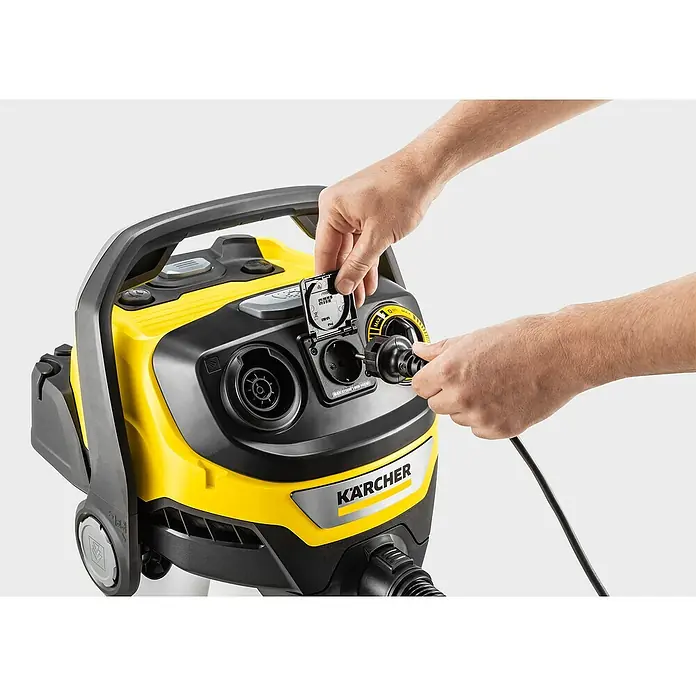Kärcher Aspirador en seco y húmedo eléctrico WD 6 P S - 1.300 W, Volumen de recipiente: 30 l - Primer plano de un aspirador de sólidos y líquidos Kärcher con una mano que conecta el cable de alimentación a la toma de corriente. El aspirador es amarillo y negro.