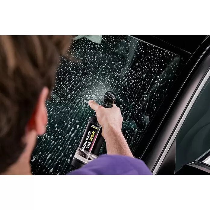 Kärcher Reinigungs-Set - 4 -tlg., Passend für: Kärcher Akku-Eiskratzer EDI 4 - Person sprüht Kärcher Autoglasreiniger aus schwarzer Flasche auf eine dunkle Autoscheibe, wobei sich weißer Schaum bildet.
