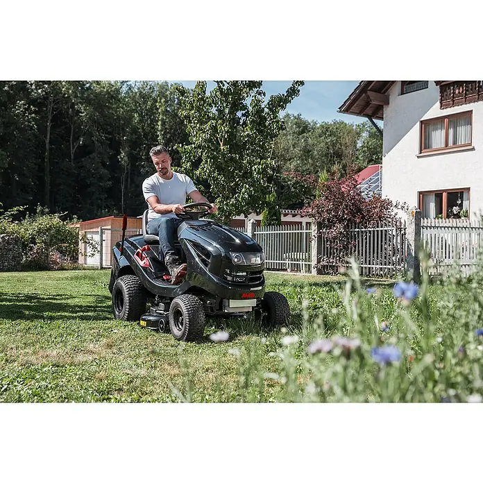 AL-KO Easy Rasentraktor T 15-93.2 HD-A - 452 cm³, Schnittbreite: 93 cm, Schnitthöhe: 80 mm - Mann fährt einen schwarzen AL-KO Aufsitzrasenmäher auf einer grünen Wiese vor einem Haus. Ideal für müheloses Rasenmähen großer Flächen.
