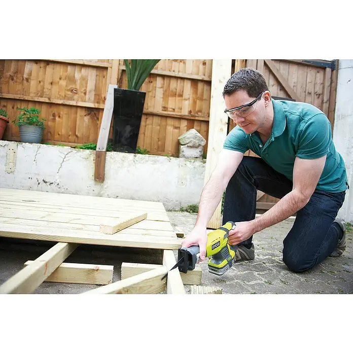 Ryobi ONE+ Maschinen-Set Combo Kit R18CK4E-242S - 18 V, 2 Akkus, 8 -tlg. - Ein Mann kniet und benutzt eine gelbe RYOBI Säbelsäge, um ein Holzstück zu schneiden, während er eine Schutzbrille trägt.